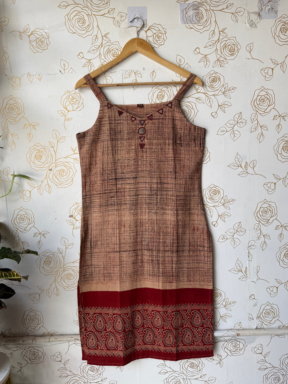 A sleeveless brown and beige patterned dress or tunic with a red paisley border, hanging on a wooden hanger against a floral wallpaper background.