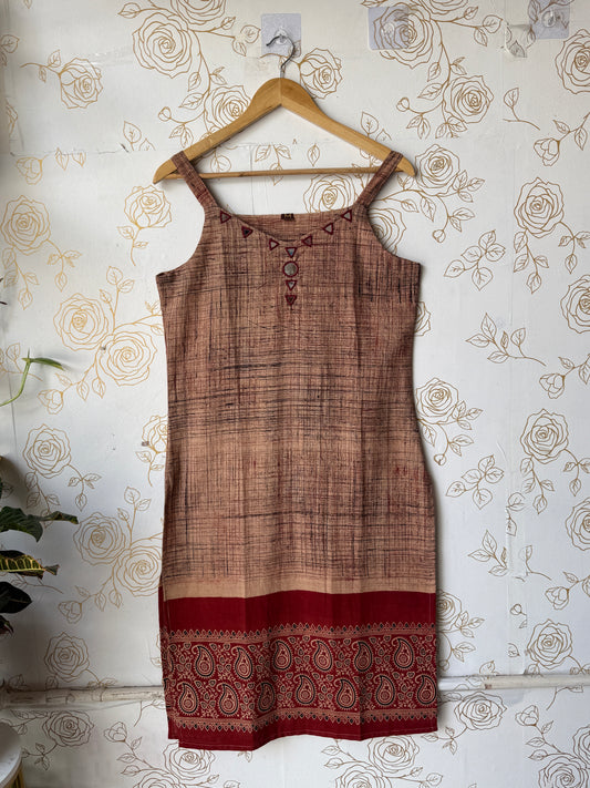 A sleeveless brown and beige patterned dress or tunic with a red paisley border, hanging on a wooden hanger against a floral wallpaper background.